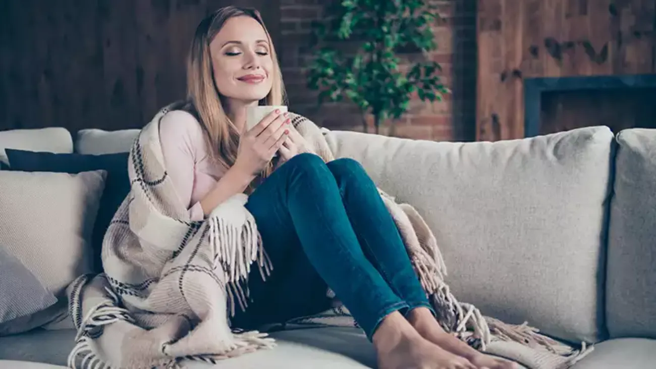A woman sitting cozily on a couch enjoying a drink out of a mug.