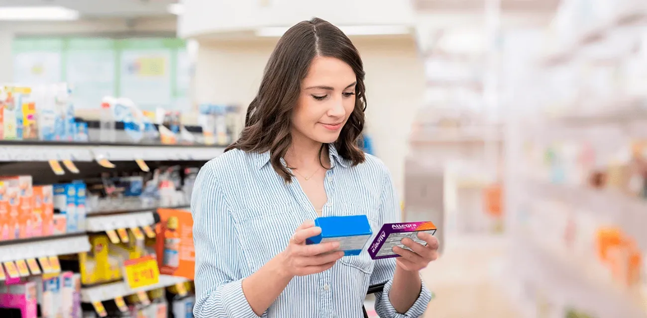 A woman comparing Allegra® with another medicine in the allergy aisle at a store, looking approvingly towards Allegra®.