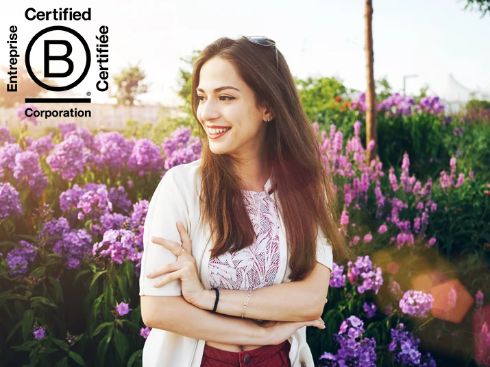 A cheerful brunette girl standing outdoors in a city park, with a backdrop of colorful flowers and bushes. Her gaze is directed playfully aside. The image includes a black B-Corp Certified logo in the upper-left corner.