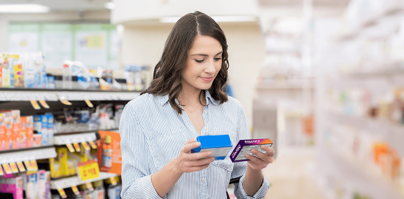 A woman comparing Allegra® with another medicine in the allergy aisle at a store, looking approvingly towards Allegra®.