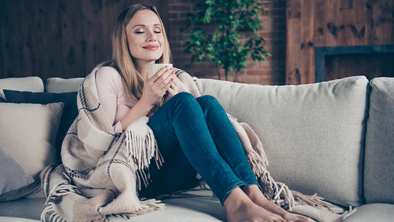 De jonge vrouw zit op de bank met een deken om zich heen en een kop koffie in haar hand te genieten.