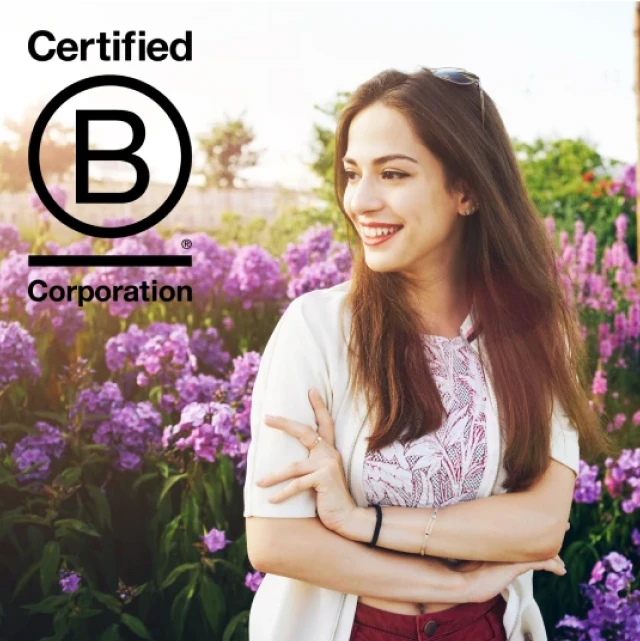 Una mujer joven sonriendo mientras se encuentra frente a un campo de flores. La imagen incluye el logo de Certified B Corporation.